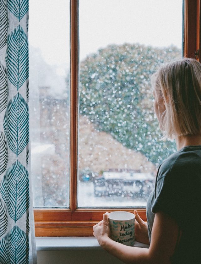 woman holding mug looking out window on dreary day looking wistful